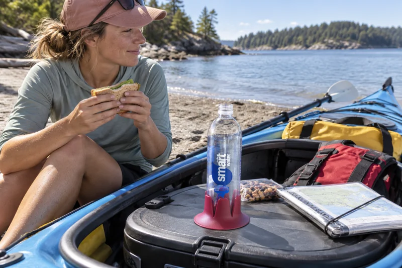 Adult seated beside beached recreational kayak during lunch stop with uncapped plastic water bottle in Steadi on wide flat dry-box lid securely level in beached cockpit at rest. AI illustration.